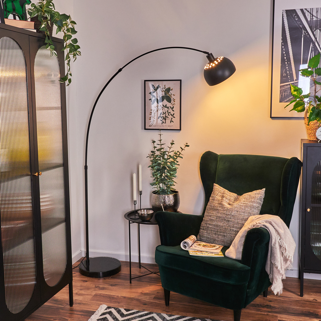 Ein dunkelgrüner Sessel mit Kissen und Decke. Eine schwarze Stehlampe, ein kleiner Tisch, eine Pflanze und ein Schrank sind ebenfalls vorhanden.