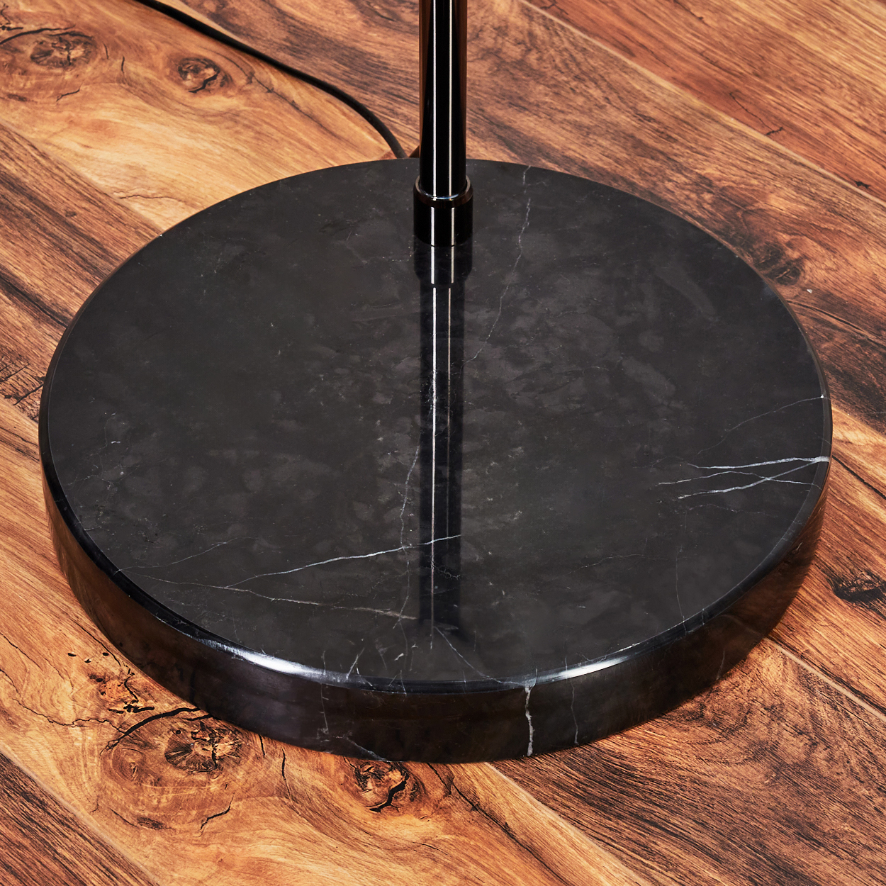 Nahaufnahme eines schwarzen Marmor-Lampenfußes auf einem Holzboden. Die Metallstange der Lampe erhebt sich aus der Mitte. Der Boden ist braun.