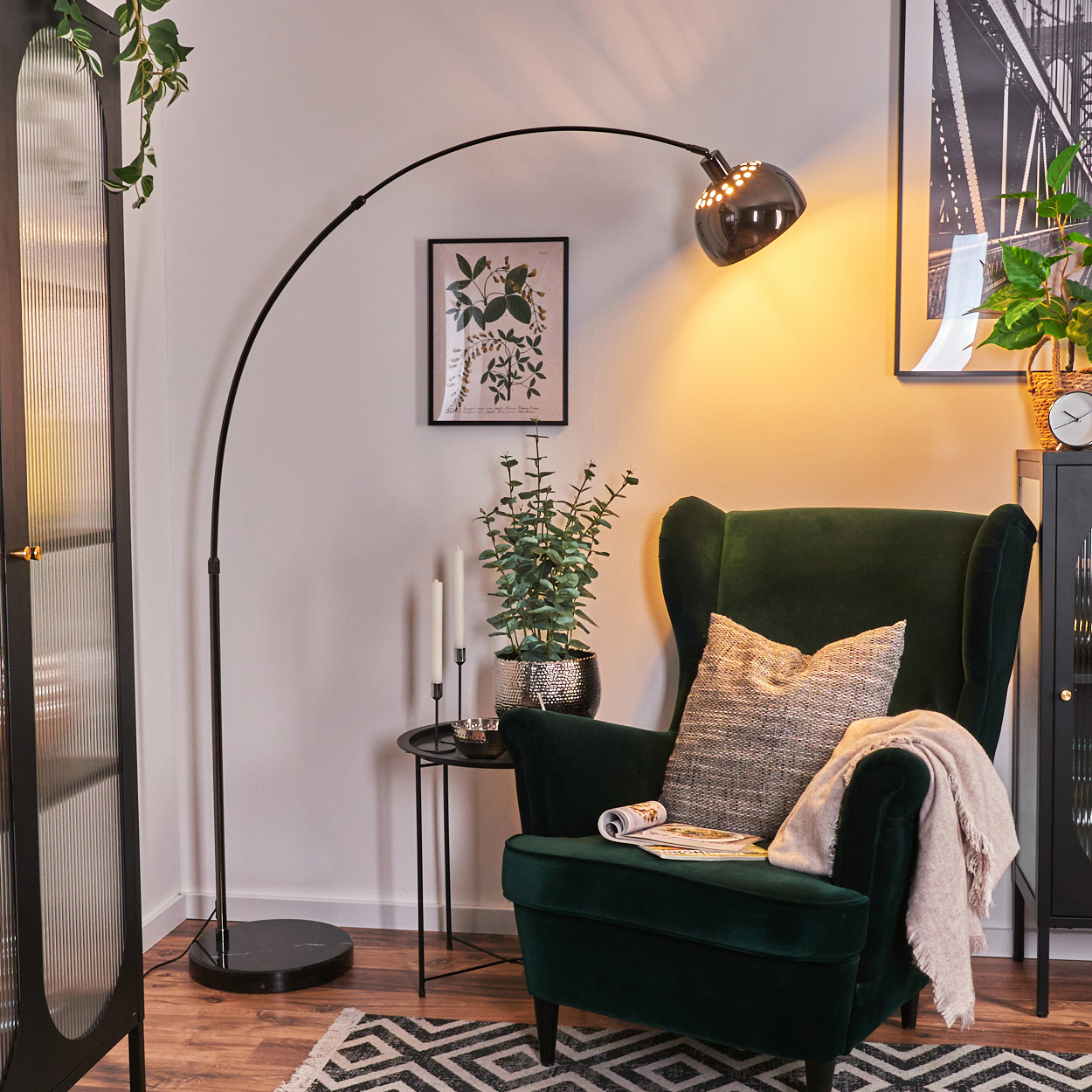 Ein grüner Sessel mit Decke und Kissen, eine Stehlampe, ein kleiner Tisch, eine Pflanze und ein Bilderrahmen.