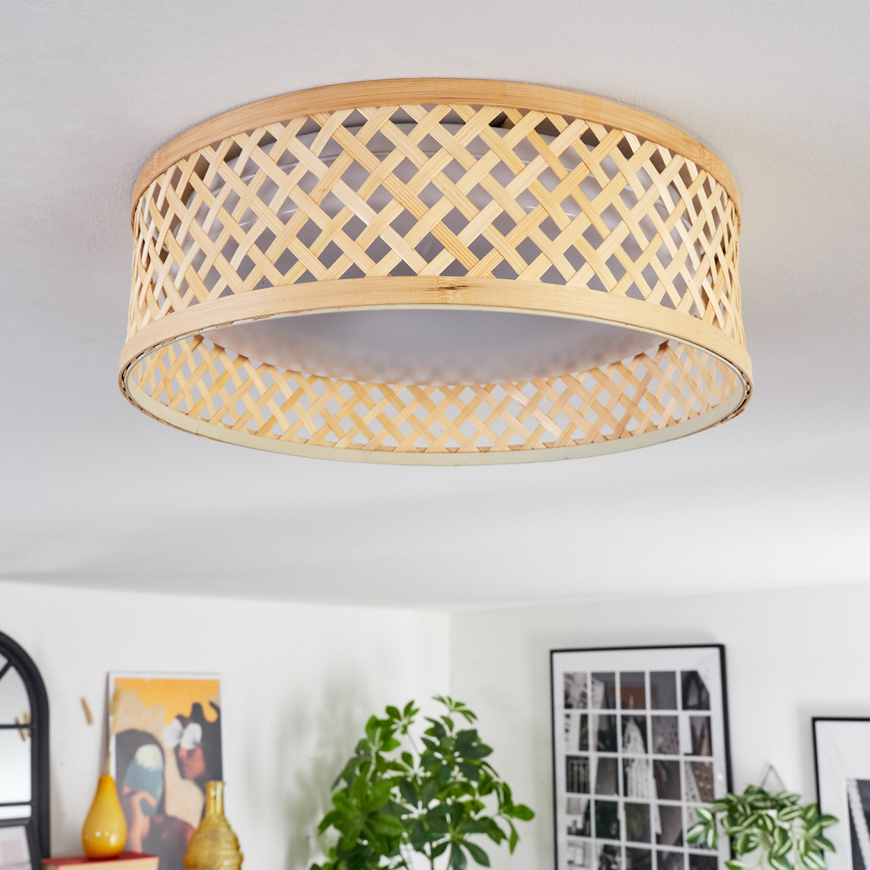 Eine Deckenlampe aus gewebtem Bambus. Die Lampe ist kreisförmig mit hellem Innenraum. Im Hintergrund befinden sich Pflanzen, Kunst und Möbel.