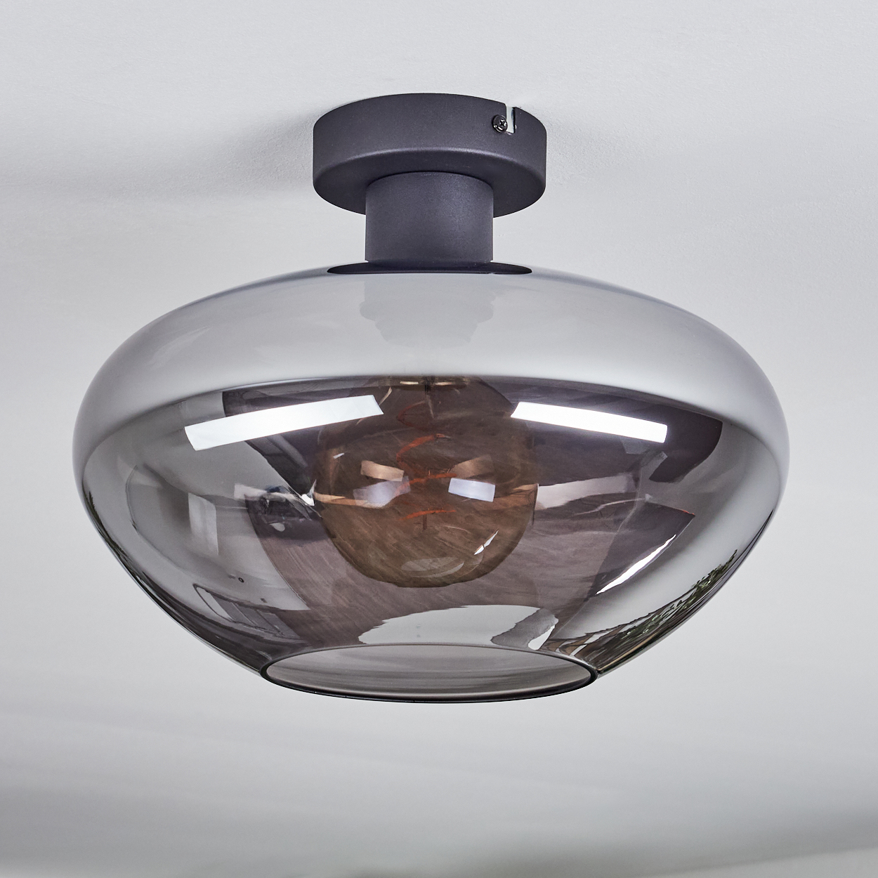 Eine moderne Deckenleuchte mit Rauchglasschirm und dunklem Metallsockel. Die Glühbirne ist im Schirm sichtbar.