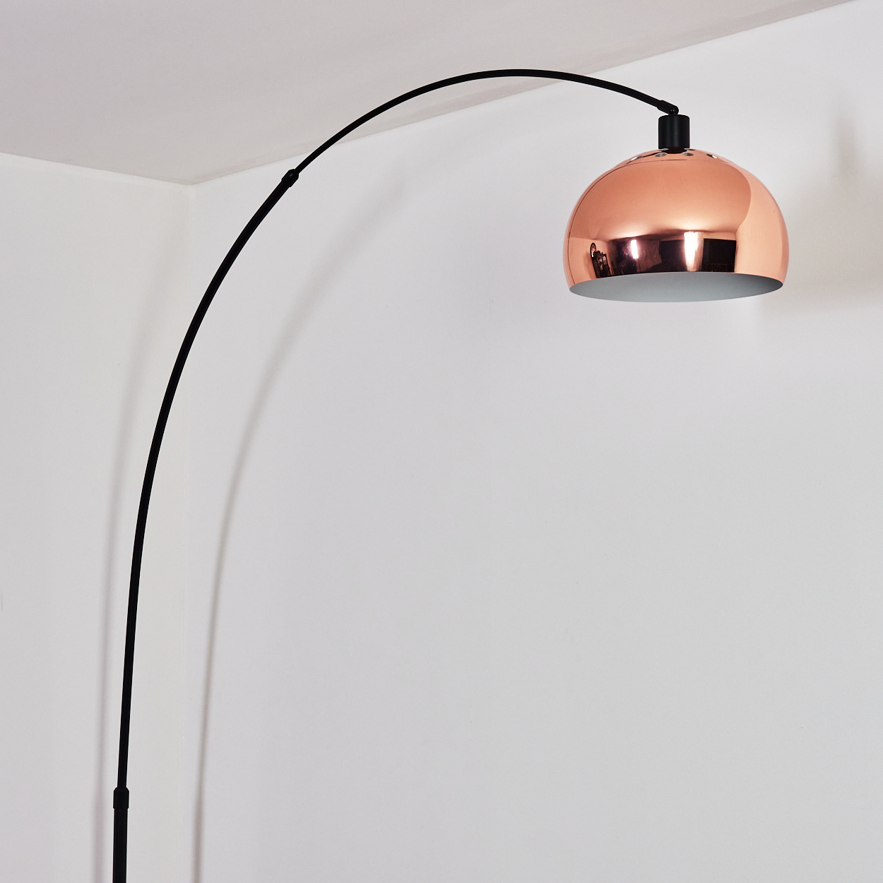 Eine moderne Bogenstehlampe mit kupferfarbenem Lampenschirm. Die Lampe hat einen schwarzen, gebogenen Arm vor einer weißen Wand.