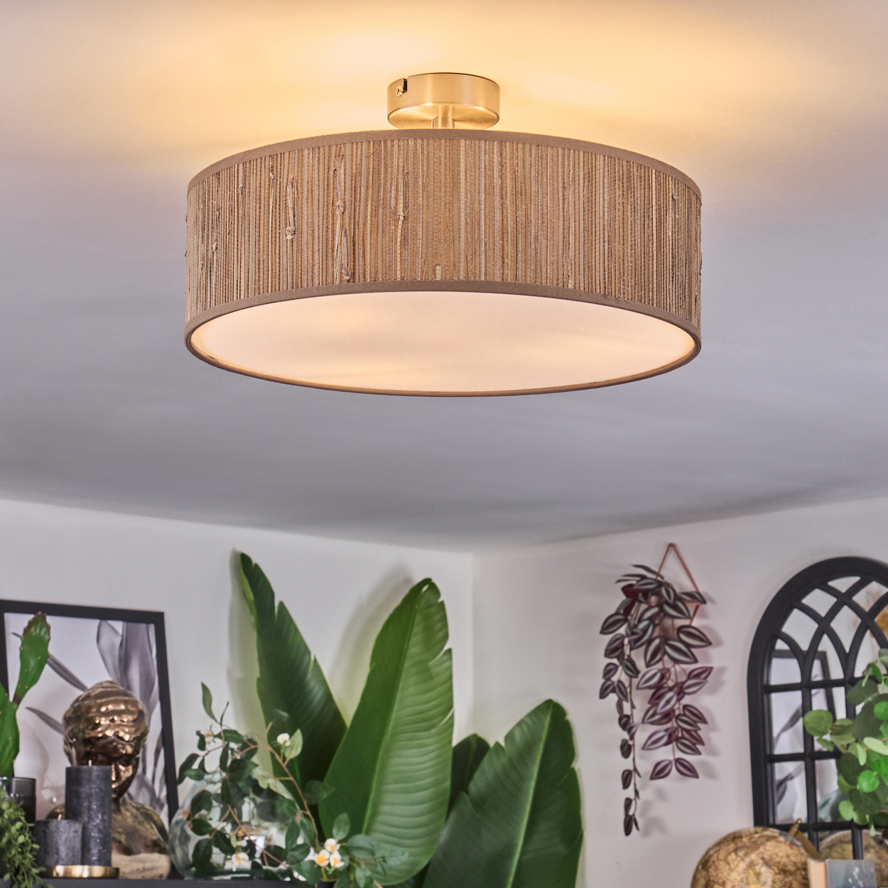 Eine Deckenleuchte mit gewebtem Lampenschirm ist an der Decke montiert. Darunter befinden sich Pflanzen und Dekorationen auf einem Regal und ein bogenförmiger Spiegel.