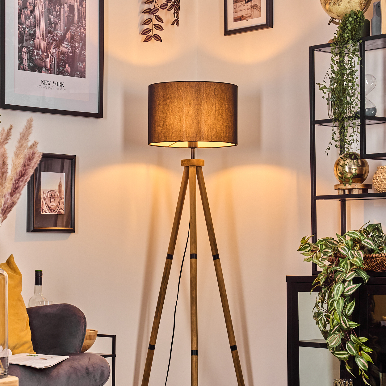 Eine Stehlampe mit schwarzem Schirm beleuchtet den Raum. Kunstwerke und Pflanzen schmücken den Raum. Im Vordergrund befindet sich ein graues Sofa.