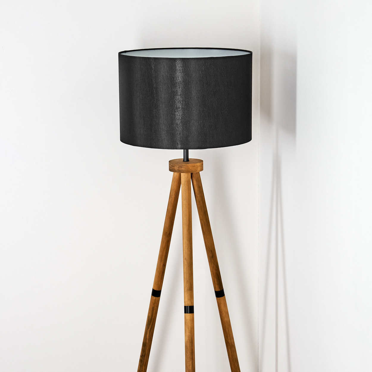 Eine hölzerne Stativlampe mit schwarzem Lampenschirm steht vor einer weißen Wand. Die Lampe wirft einen Schatten.