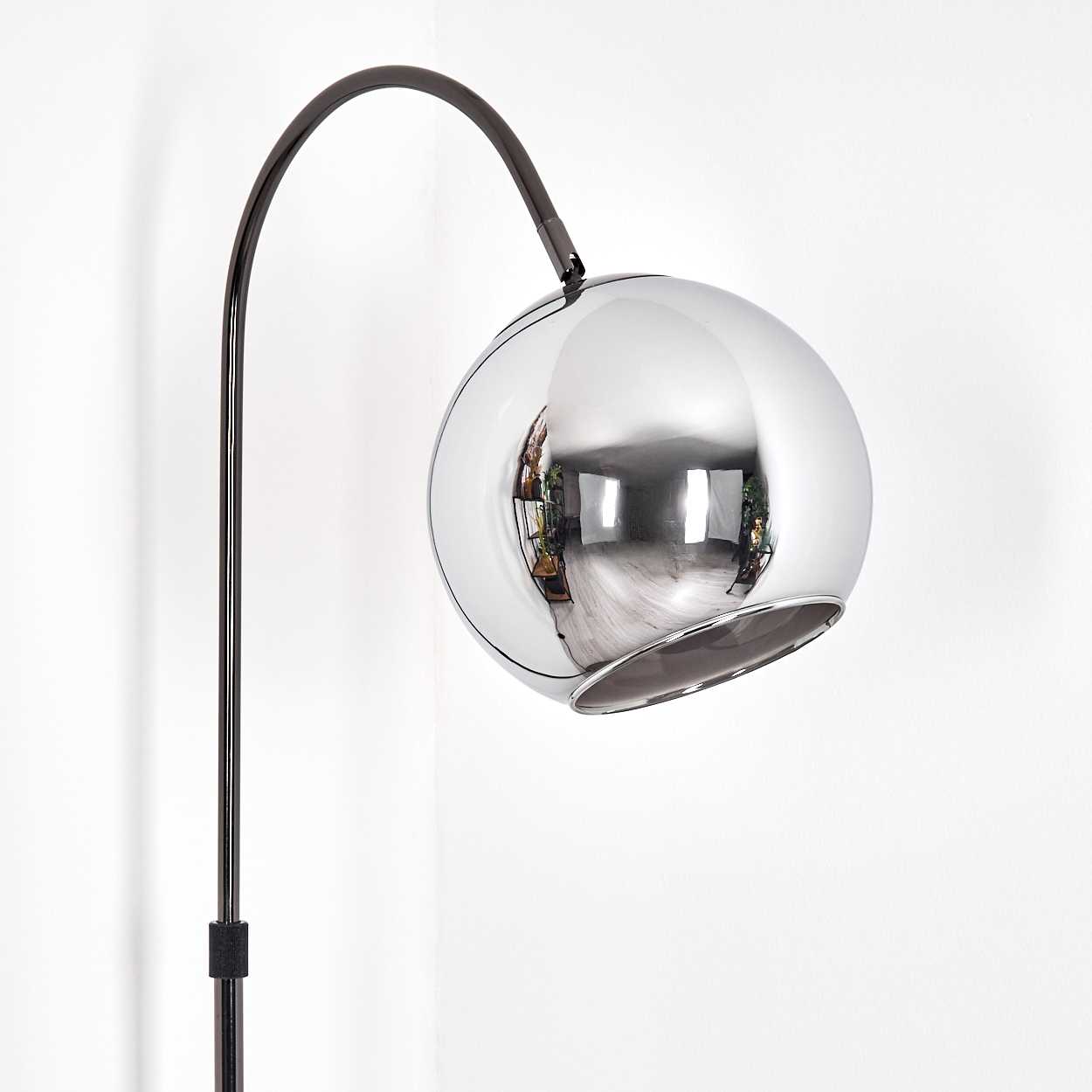 Eine verchromte Kugellampe reflektiert einen Raum mit Fenstern und Pflanzen. Ein schwarzer Ständer stützt die Lampe an einer weißen Wand.
