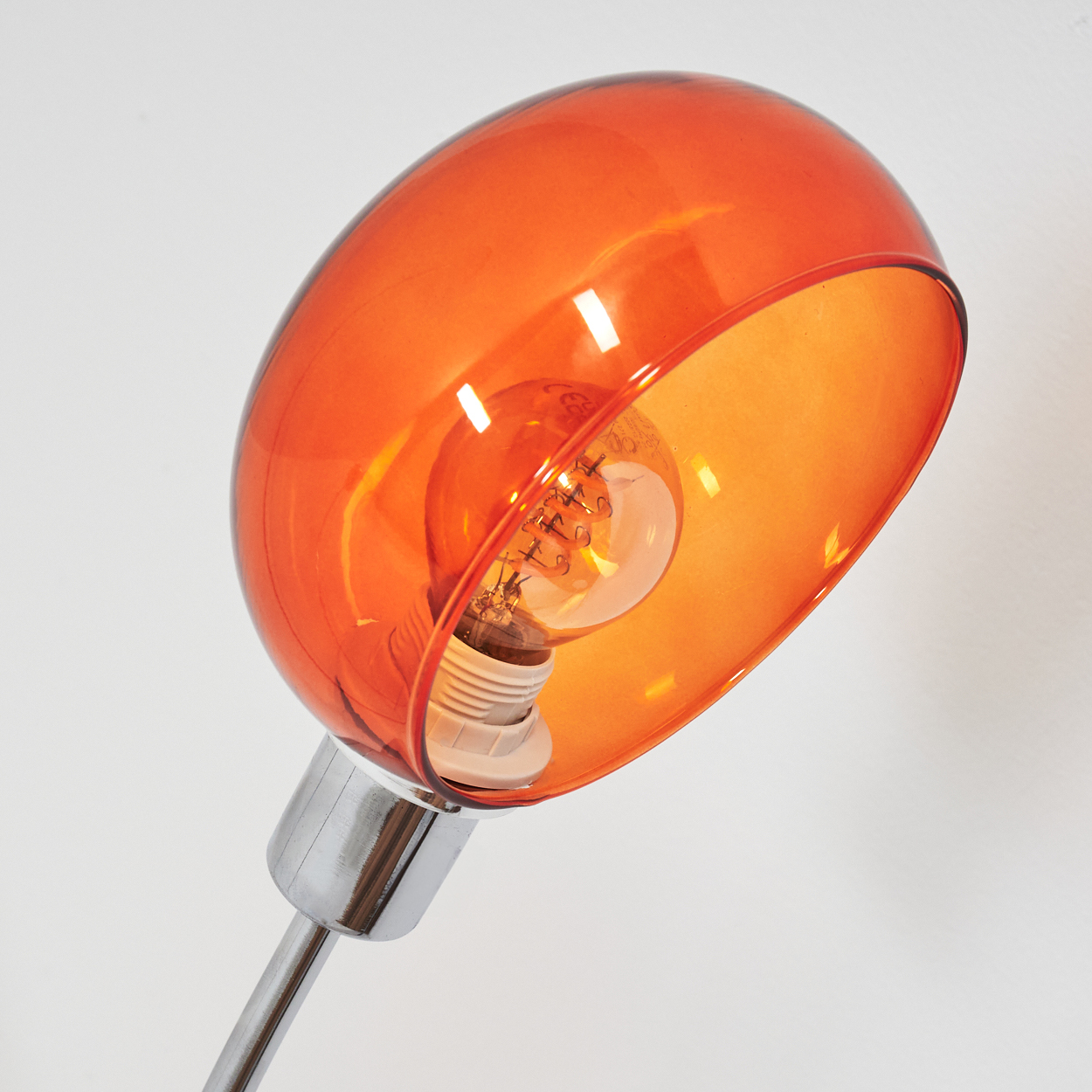 Eine orangefarbene Lampe mit verchromtem Sockel. Die Glühbirne ist im orangenen Schirm vor weißem Hintergrund sichtbar.