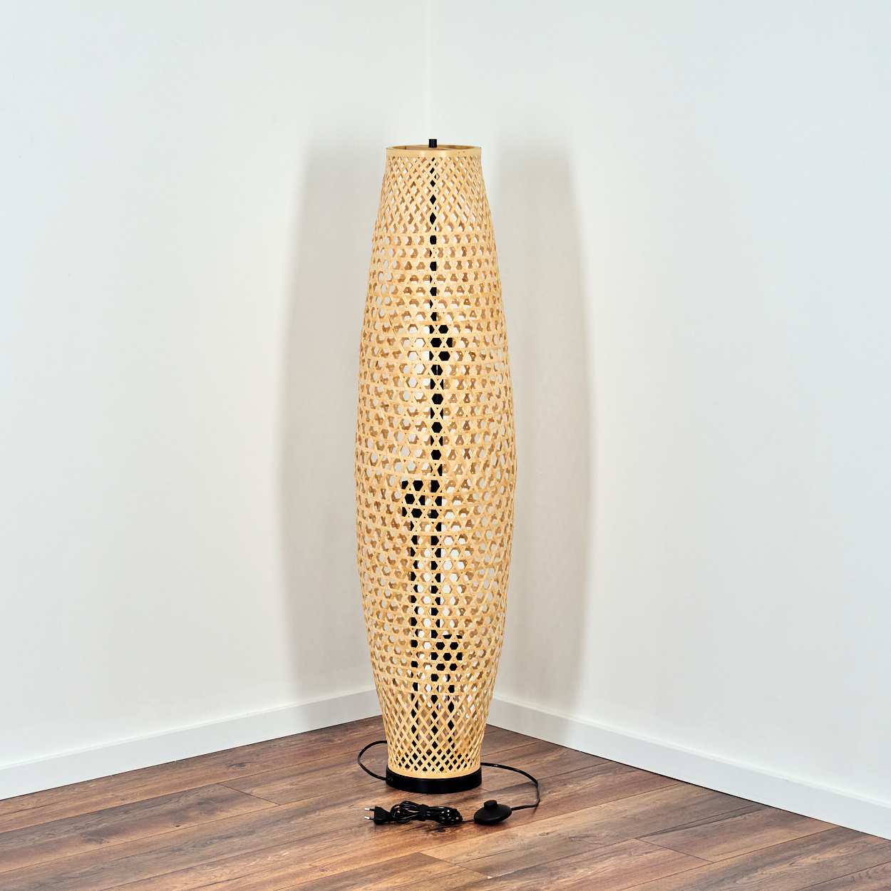 Eine gewebte Bambus-Stehlampe steht in einer Ecke, mit schwarzem Sockel und Kabel. Weiße Wände und Holzböden bilden den Hintergrund.