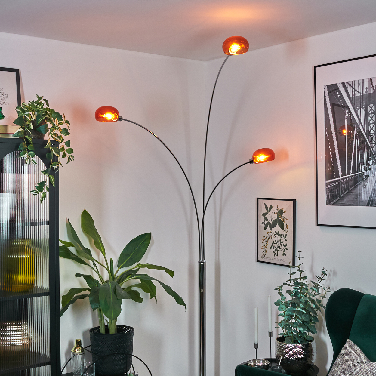 Ein modernes Wohnzimmer mit Stehlampe. Die Lampe hat drei orangefarbene Kugeln. Pflanzen und gerahmte Bilder schmücken die weißen Wände.