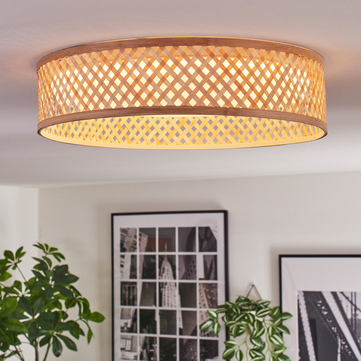 Eine runde, gewebte Deckenlampe aus Bambus. Darunter befinden sich gerahmte Bilder und Pflanzen. Die Lampe ist beleuchtet.