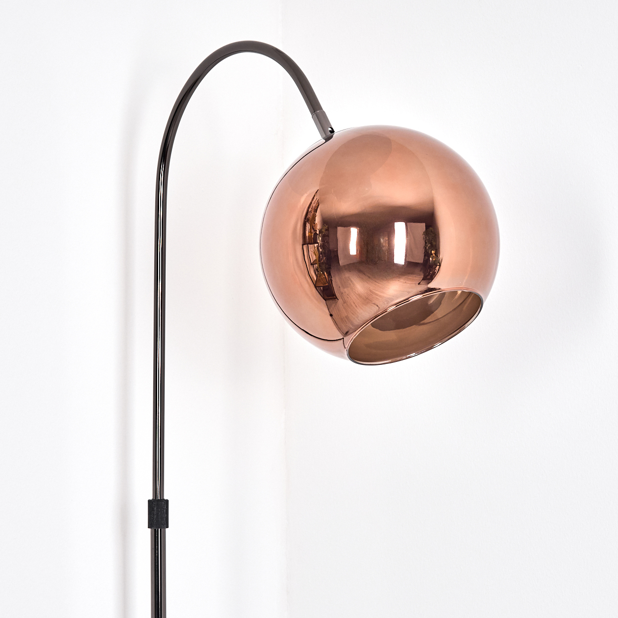 Eine moderne Stehlampe mit kupferfarbenem Kugelschirm und schwarzem, gebogenem Arm steht in einer Ecke an einer weißen Wand.