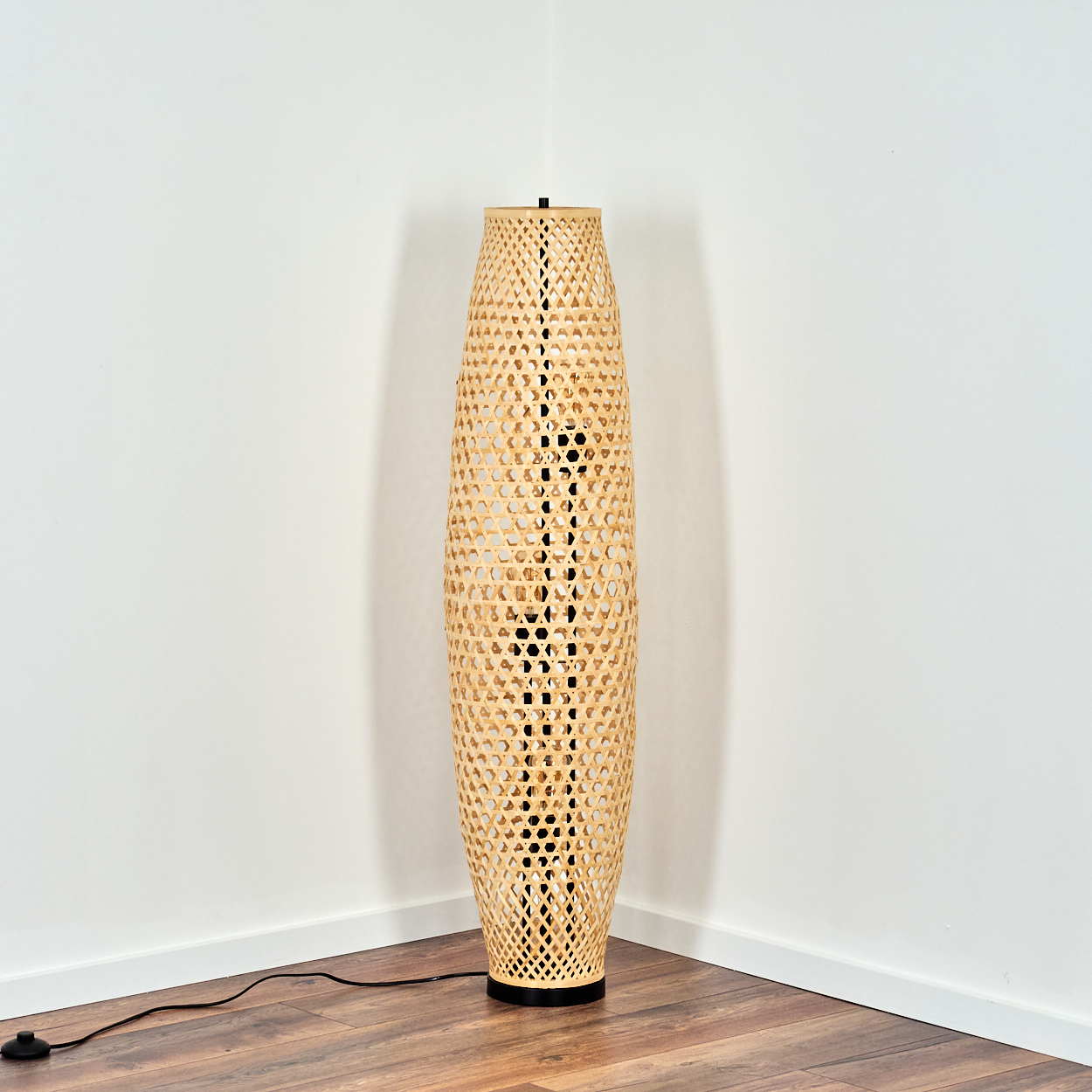 Eine hohe, gewebte Stehlampe aus Bambus steht in einer Ecke. Sie hat einen schwarzen Sockel und ein Netzkabel. Der Boden ist aus Holz.