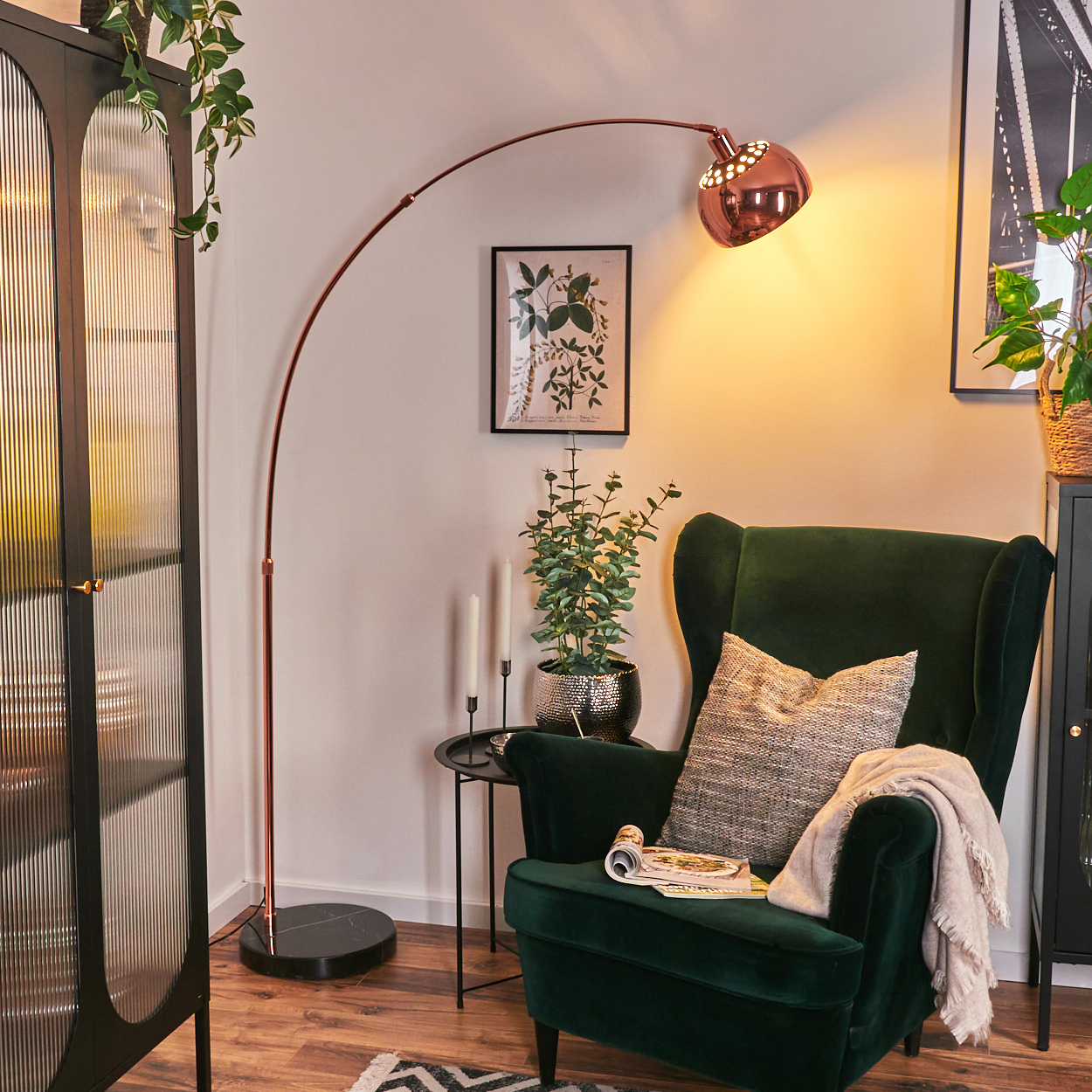 Ein Wohnzimmer mit grünem Sessel, kupferfarbener Stehlampe und schwarzem Schrank mit Glastüren. Ein Blumentopf steht auf einem Beistelltisch.