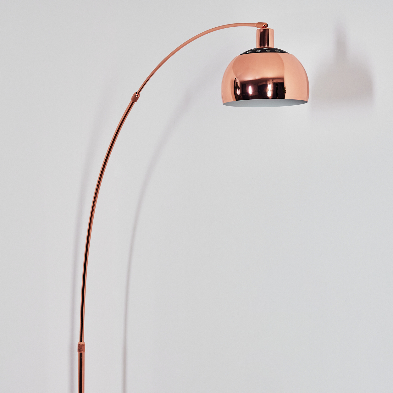 Eine kupferfarbene Bogenstehlampe steht vor einer weißen Wand. Die Lampe hat einen großen, kuppelförmigen Schirm.
