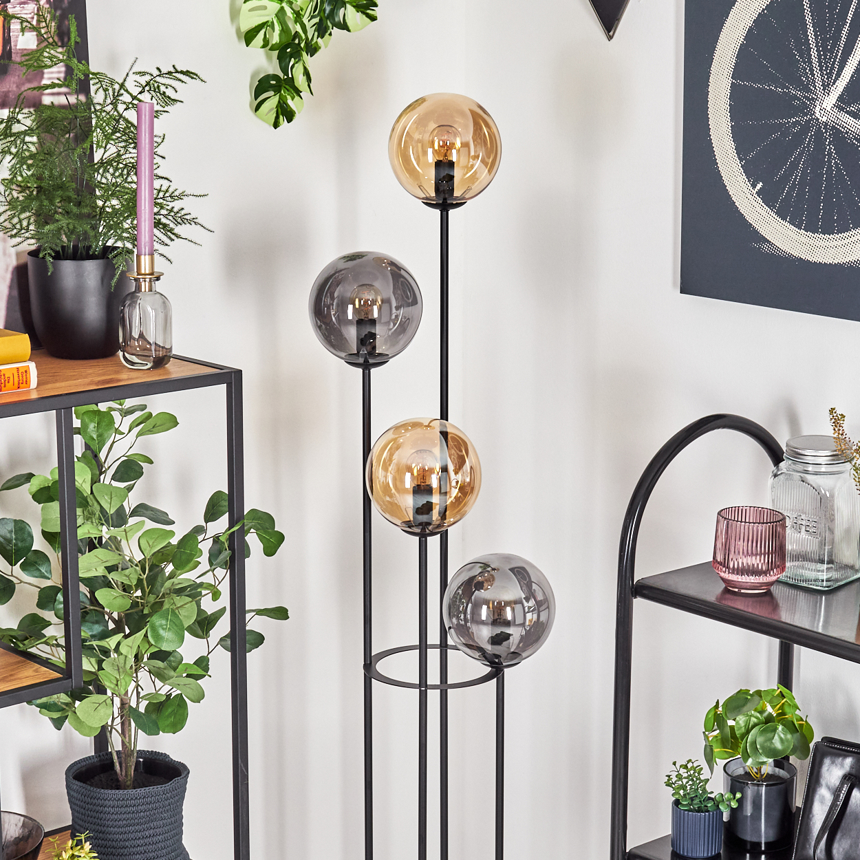 Eine moderne Stehlampe mit vier Glaskugeln, Pflanzen und Regalen vor einer weißen Wand. Die Lampe hat bernsteinfarbene und graue Kugeln.