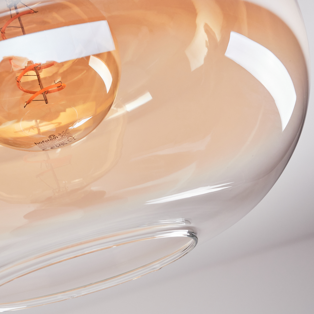 Nahaufnahme einer Glasleuchte mit einer bernsteinfarbenen Glühbirne. Das Glas hat eine gebogene Form mit klarer, reflektierender Oberfläche.