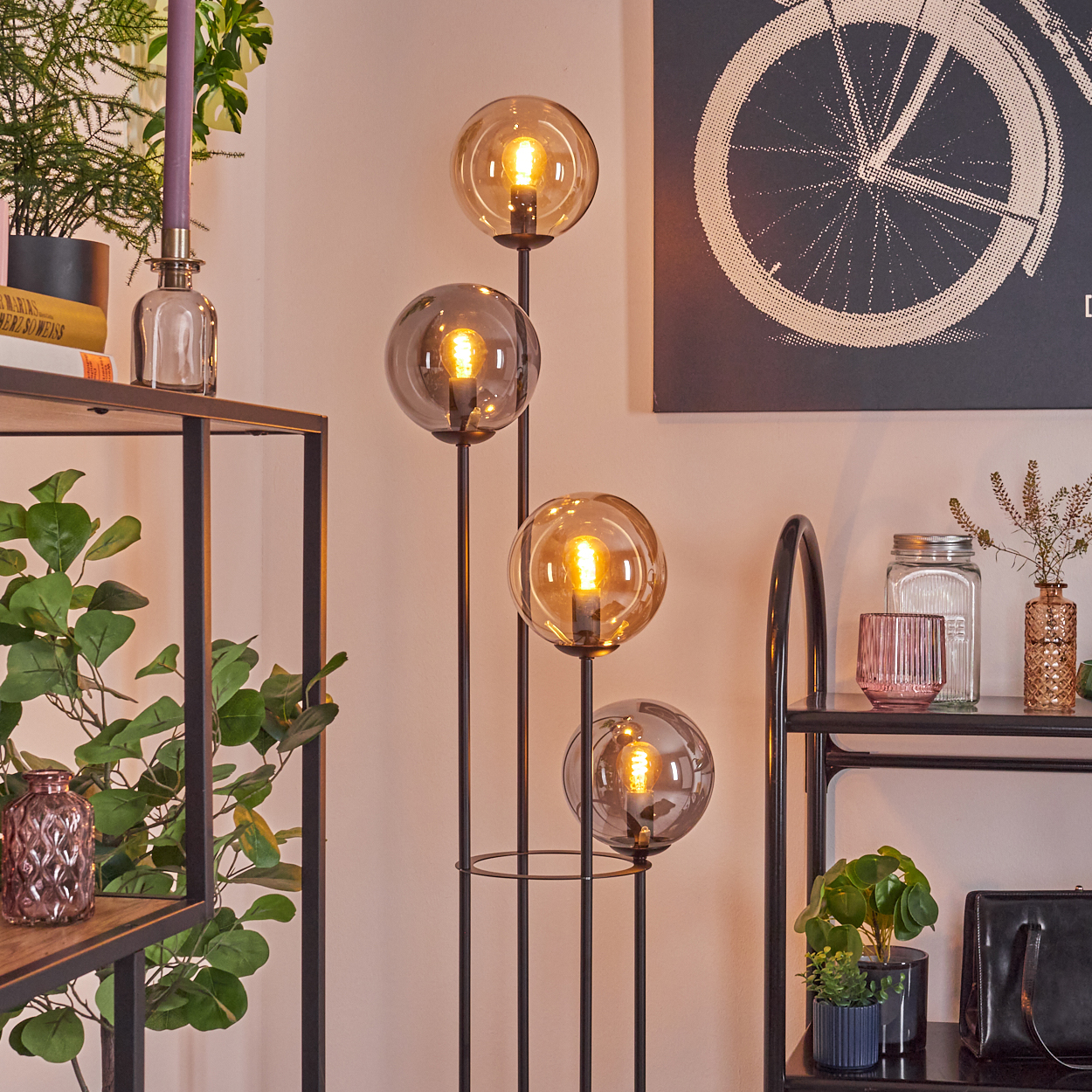 Eine Stehlampe mit vier leuchtenden Kugeln steht in der Nähe von Regalen mit Pflanzen und Dekoration. Ein Fahrradrad-Kunstwerk hängt an der Wand.
