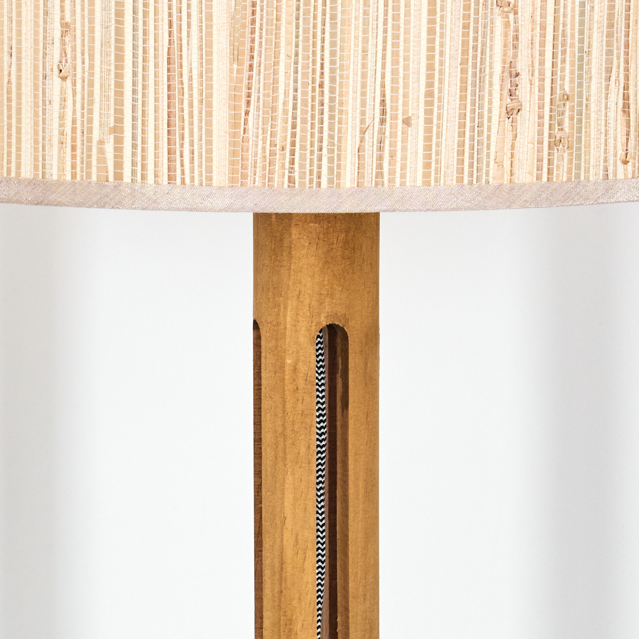 Nahaufnahme einer Lampe. Der Lampenschirm ist aus beigem Stoff gewebt. Der Holzsockel hat Aussparungen, die ein schwarz-weißes Kabel freilegen.