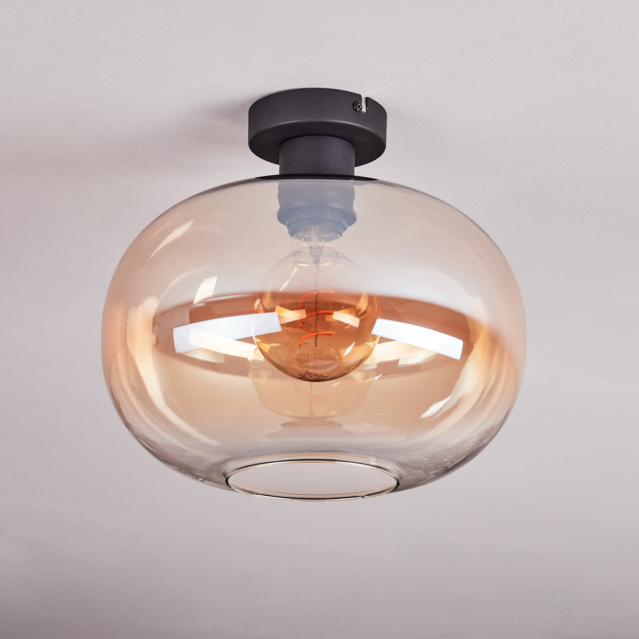 Eine moderne Deckenleuchte. Sie hat einen runden, bernsteinfarbenen Glasschirm mit einer sichtbaren Glühbirne. Sie ist auf einem dunklen Sockel montiert.