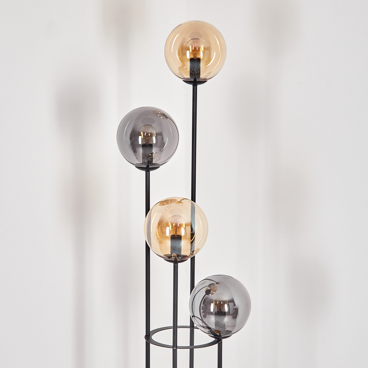 Moderne Stehlampe mit vier Glaskugelschirmen in Bernstein und Grau. Schwarzes Metallgestell. Weiße Wand im Hintergrund.