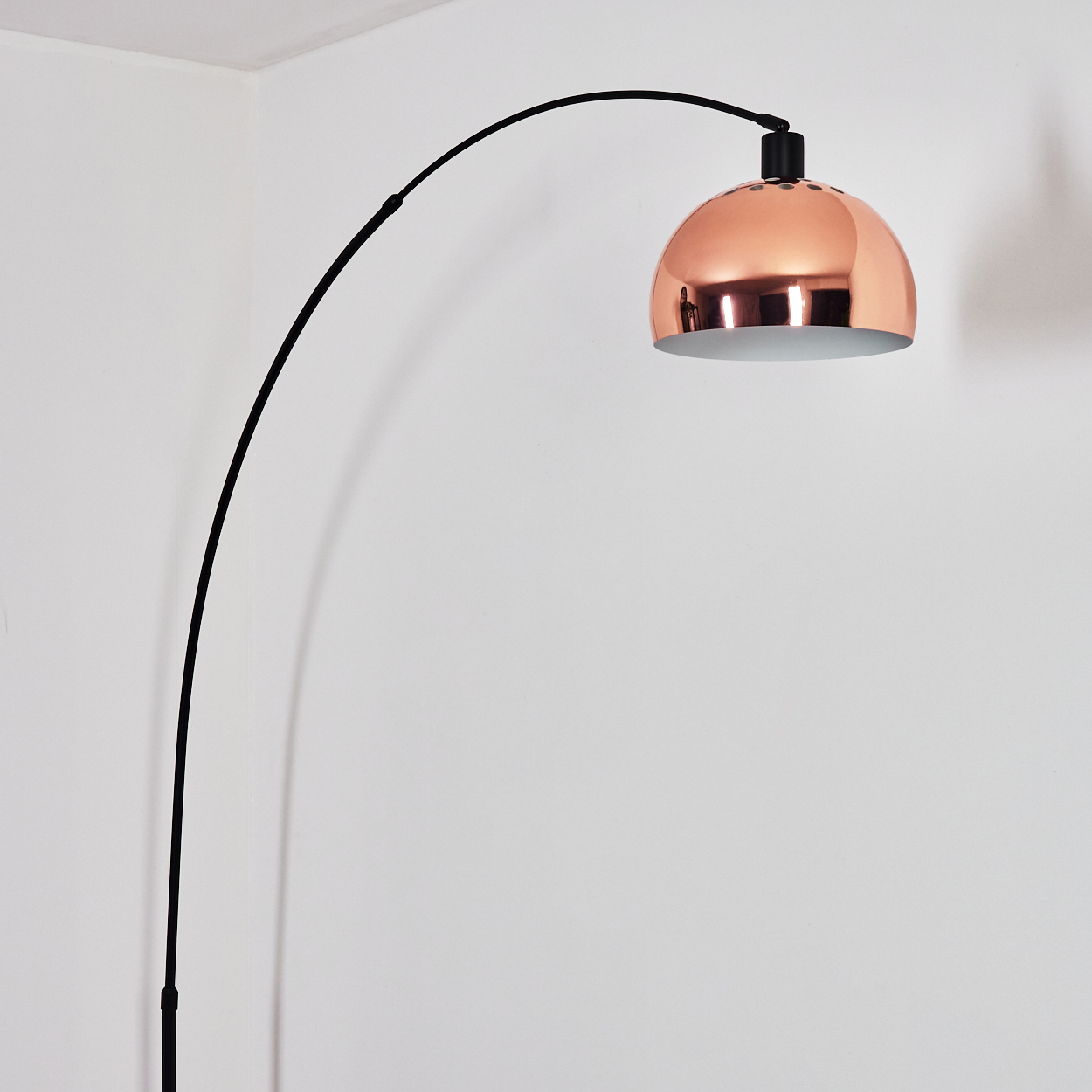 Eine moderne Bogenlampe mit kupferfarbenem Lampenschirm. Die Lampe hat einen schwarzen, gebogenen Arm und steht vor einer weißen Wand.