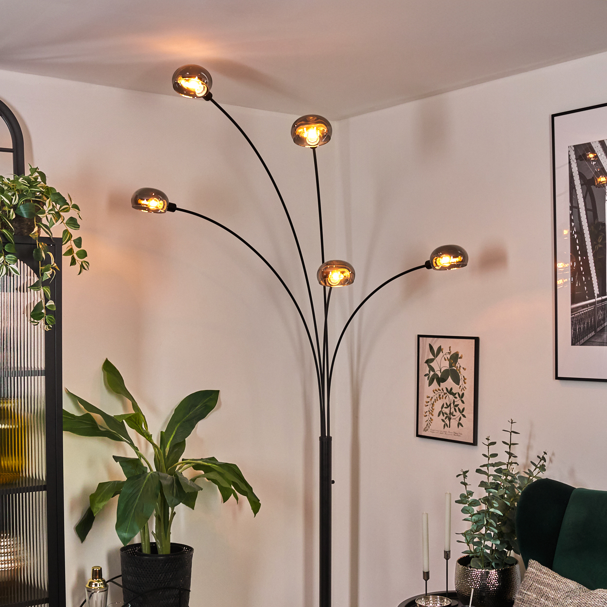 Ein modernes Wohnzimmer mit einer schwarzen Stehlampe mit fünf Glühbirnen. Es gibt Pflanzen, ein gerahmtes Bild und einen grünen Samtstuhl.
