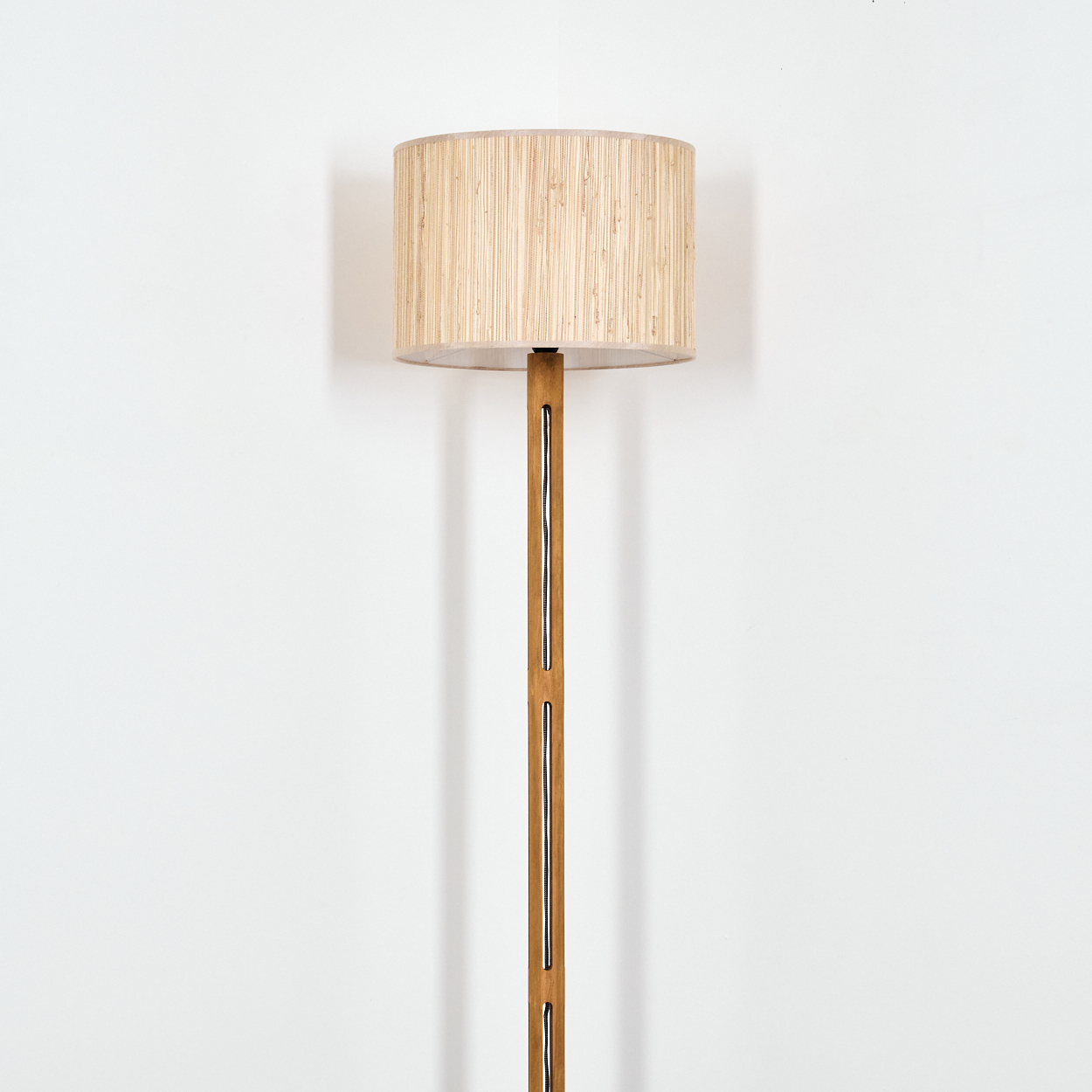 Eine Stehlampe mit beigem Lampenschirm und Holzstange. Der Hintergrund ist eine schlichte weiße Wand.