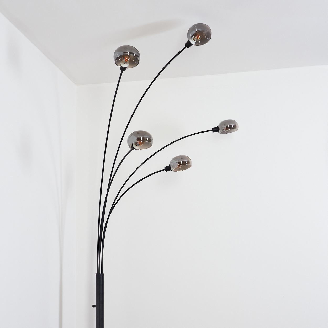Eine moderne Stehlampe mit fünf gebogenen Armen, die jeweils einen rauchigen Glaskolben halten. Die Lampe ist schwarz, vor weißen Wänden.