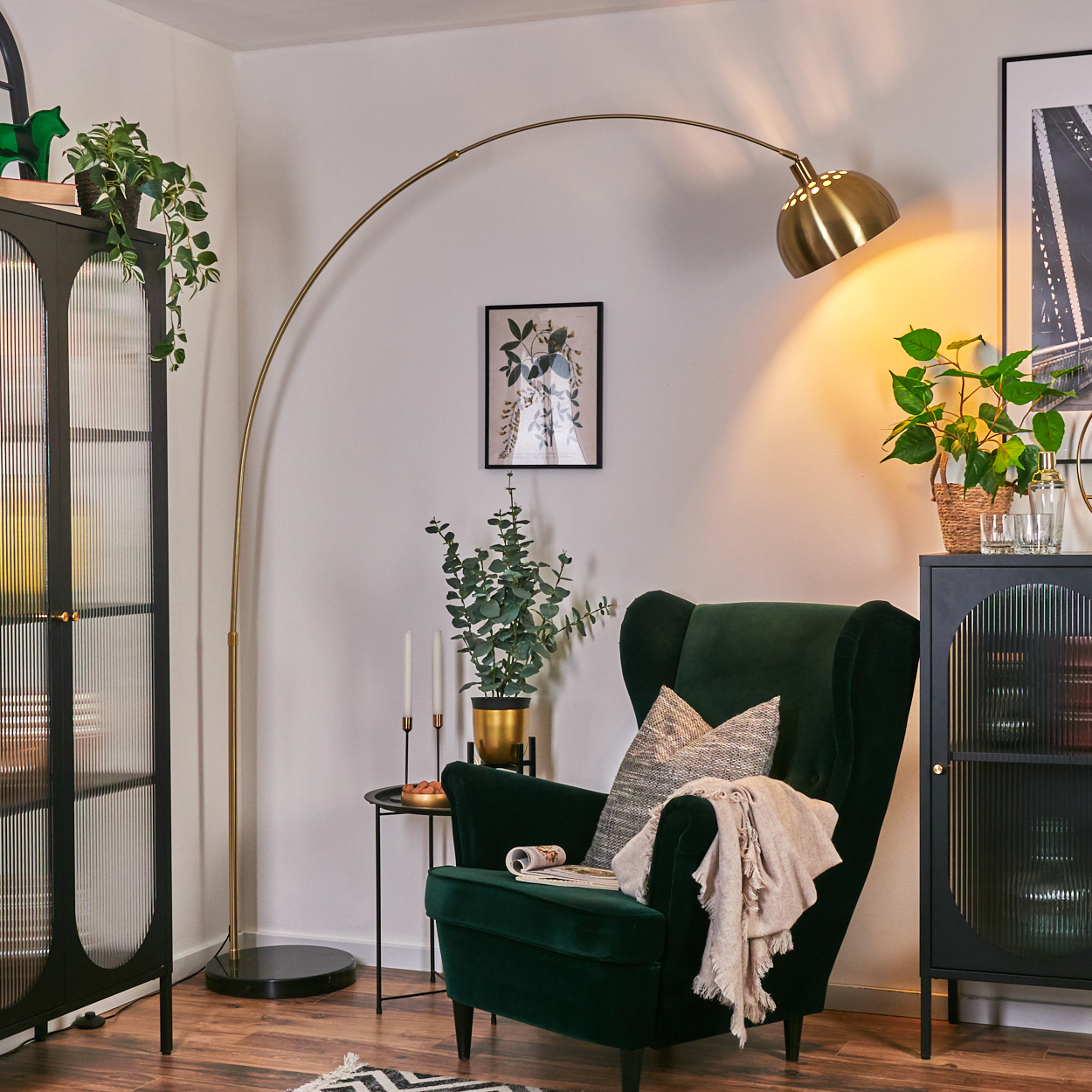 Ein grüner Samt-Sessel mit Decke und Kissen, eine goldene Stehlampe, schwarze Schränke und Topfpflanzen in einem Wohnzimmer.