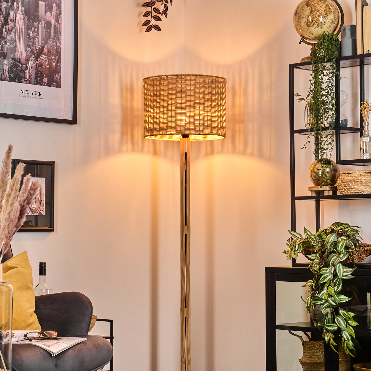 Eine beleuchtete gewebte Lampe steht in einem Raum. Ein schwarzes Regal mit Pflanzen und Globus befindet sich rechts. Links ist ein gerahmtes New York-Bild.