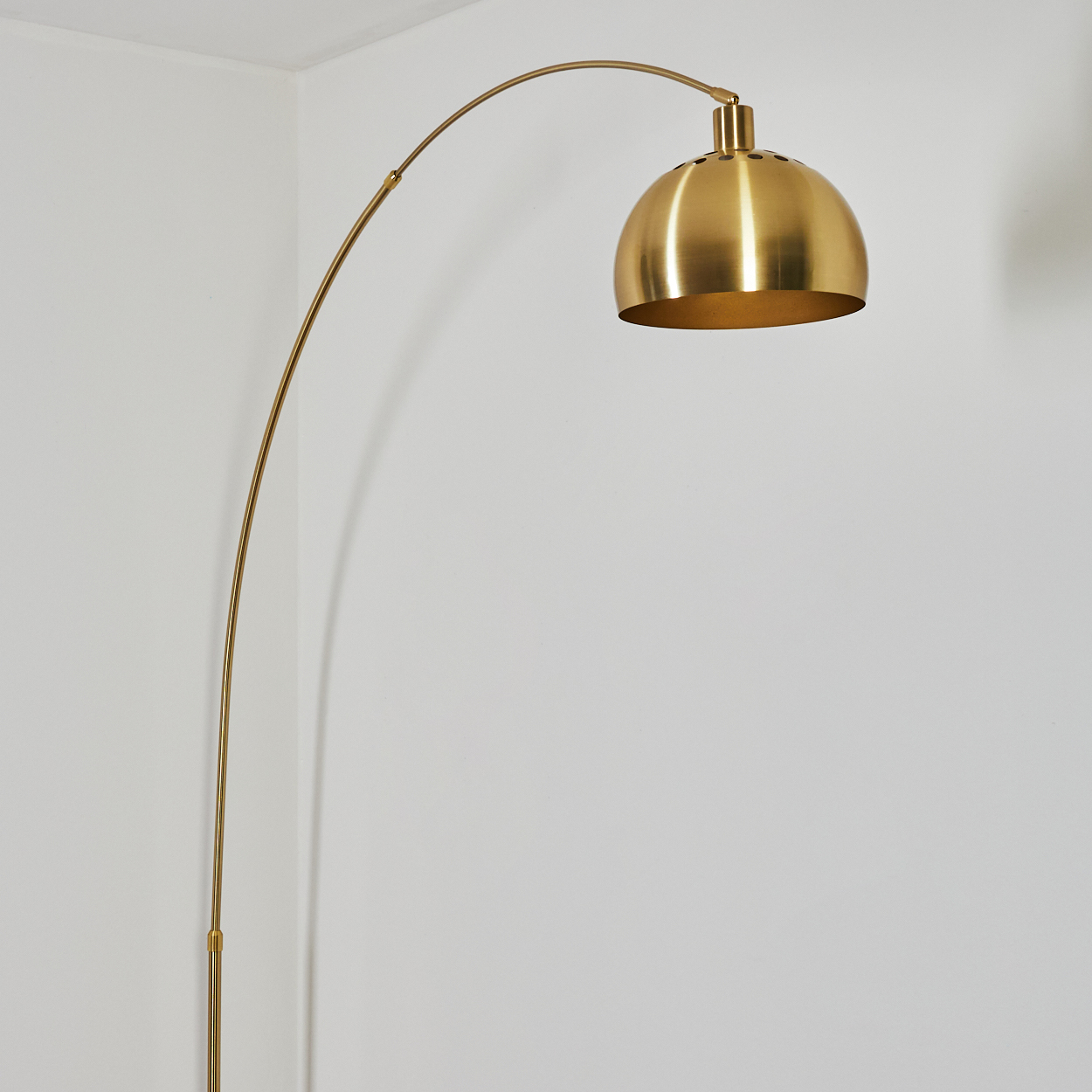 Eine goldene Bogen-Stehlampe steht vor einer weißen Wand. Die Lampe hat einen kuppelförmigen Schirm und einen gebogenen Schaft.