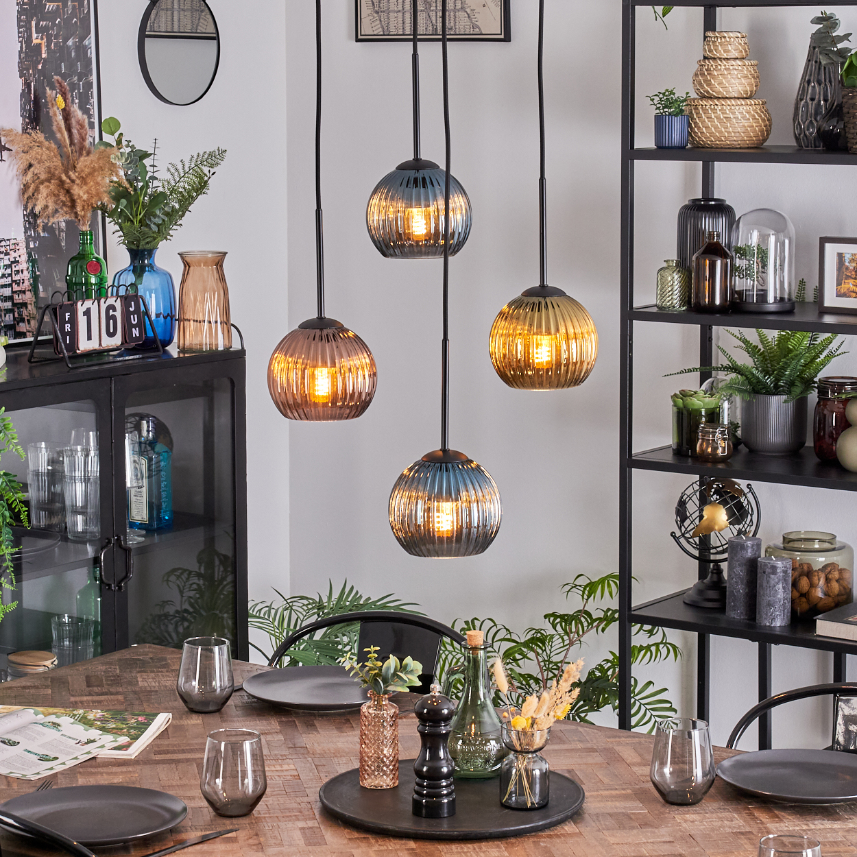 Esszimmer mit Holztisch, Hängelampen und einem schwarzen Regal. Tischdekoration umfasst Gläser und Teller. Dekorative Gegenstände.