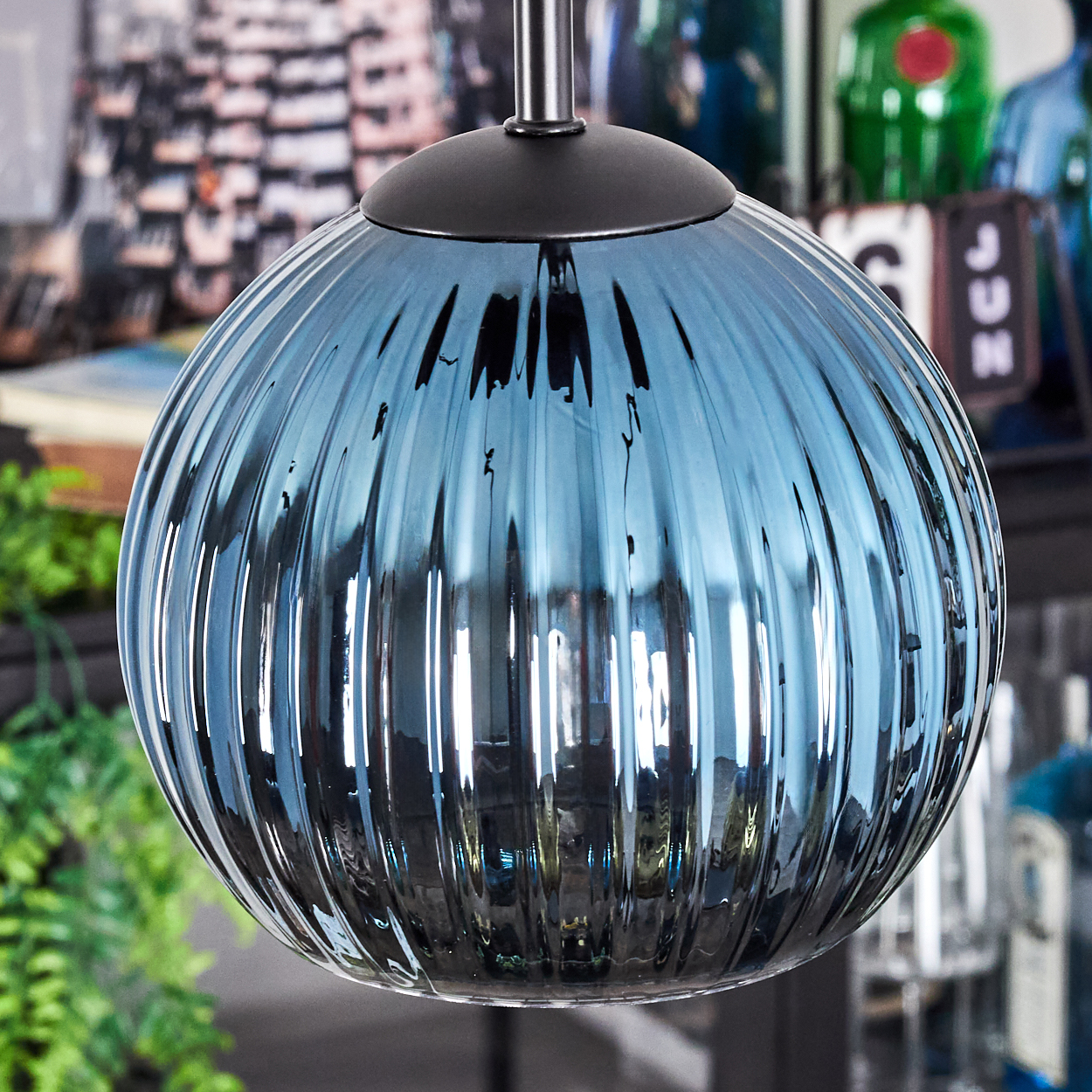 Eine runde, blaue Glas-Pendelleuchte hängt an einer schwarzen Halterung. Das Glas ist gerippt. Hintergrund: verschwommene Dekoration und grüne Pflanzen.
