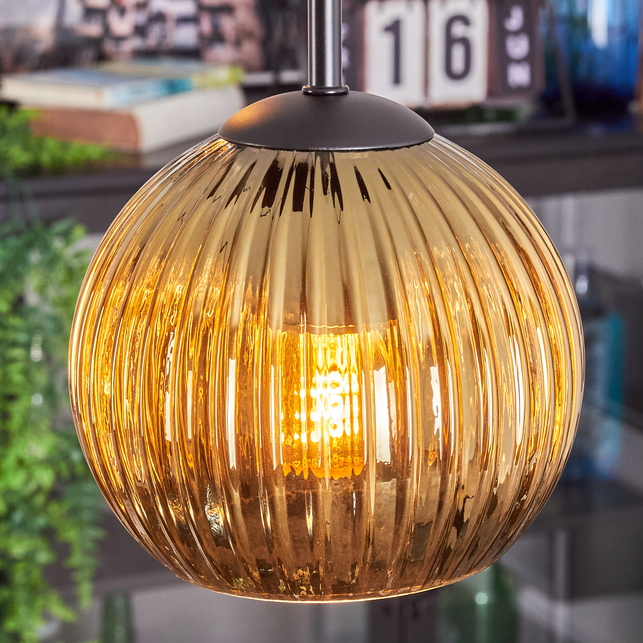Eine goldene, gerippte Glas-Pendelleuchte mit einer leuchtenden Glühbirne. Der Hintergrund zeigt verschwommenes Dekor und eine Kalenderseite mit der Zahl 16.
