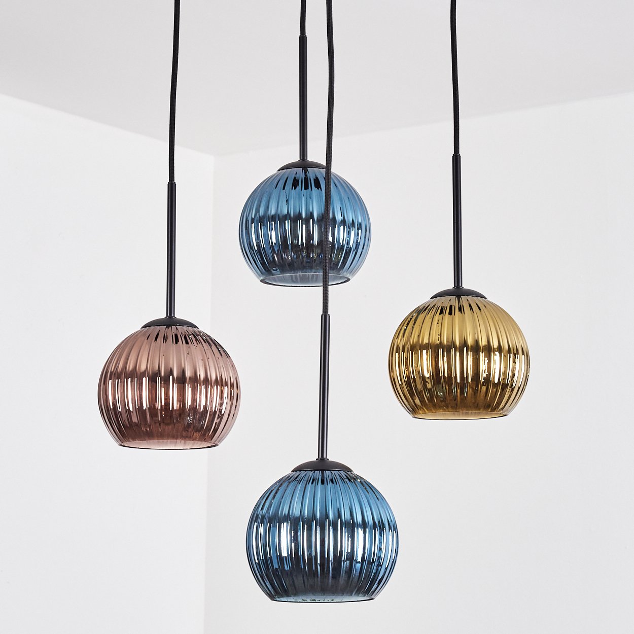 Vier bunte Pendelleuchten. Eine rosa, zwei blaue und eine goldene. Alle sind kugelförmig mit schwarzen Kabeln und vor einer weißen Wand.