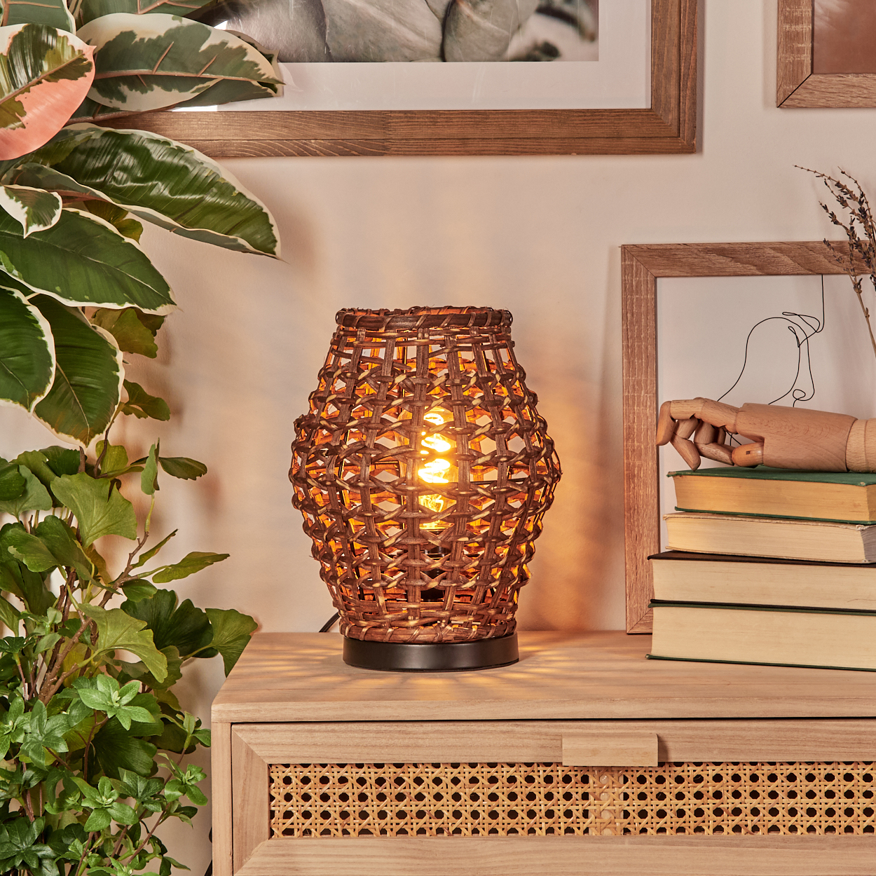 Eine gewebte Lampe mit einer brennenden Glühbirne steht auf einem Holzschrank. Pflanzen und gerahmte Kunstwerke sind im Hintergrund.