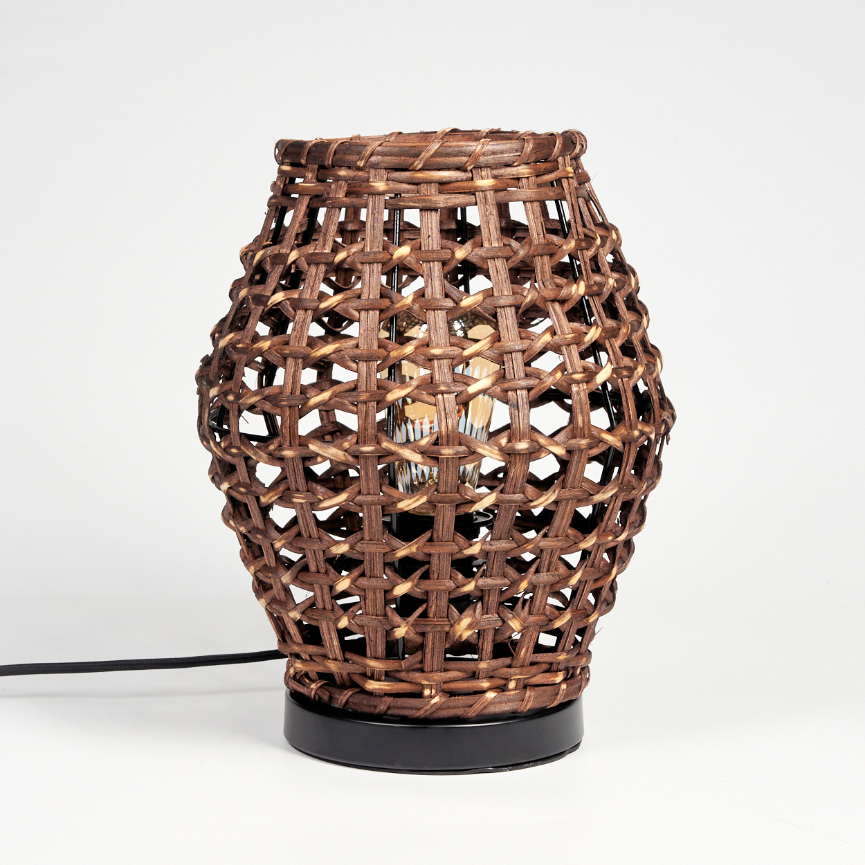 Eine braune Lampe mit einem Webmuster und einer Glühbirne im Inneren, auf einem schwarzen Sockel. Ein schwarzes Kabel erstreckt sich.