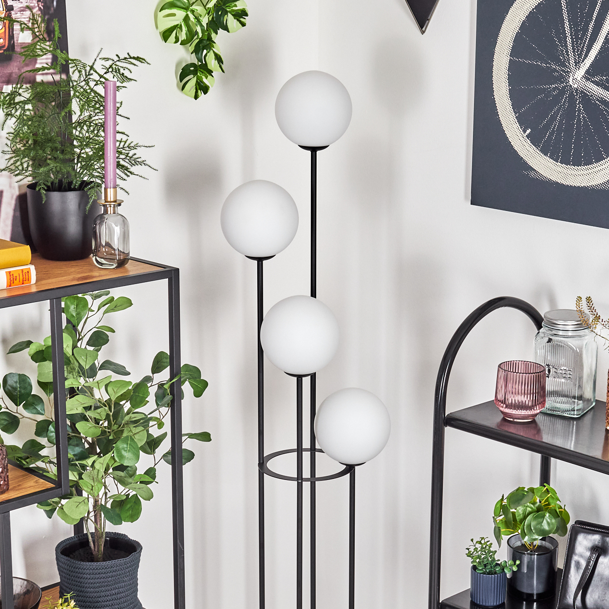 Eine moderne Stehlampe mit vier weißen Kugeln steht neben Regalen mit Pflanzen und Dekoration. Ein Fahrradrad-Kunstwerk ist sichtbar.