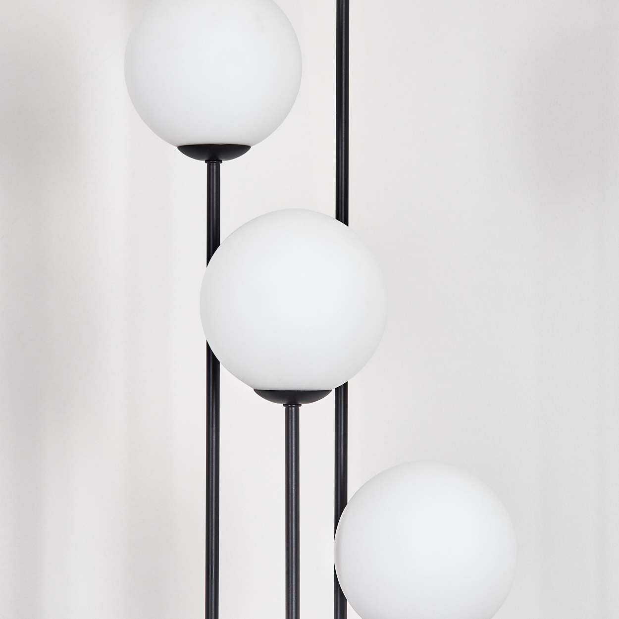 Eine moderne Lampe mit drei weißen Kugelleuchten steht vor einer weißen Wand. Die Lampe hat schwarze Stangen.