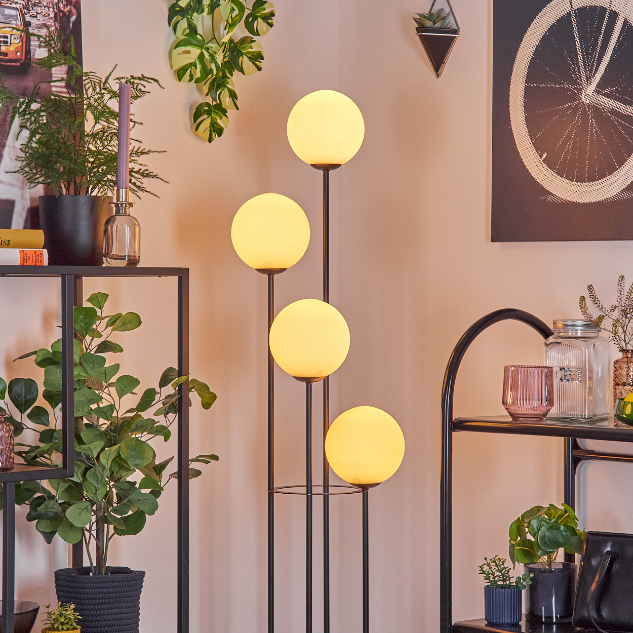 Ein modernes Wohnzimmer mit einer hohen Lampe mit vier leuchtenden Kugeln, umgeben von Pflanzen und Regalen.