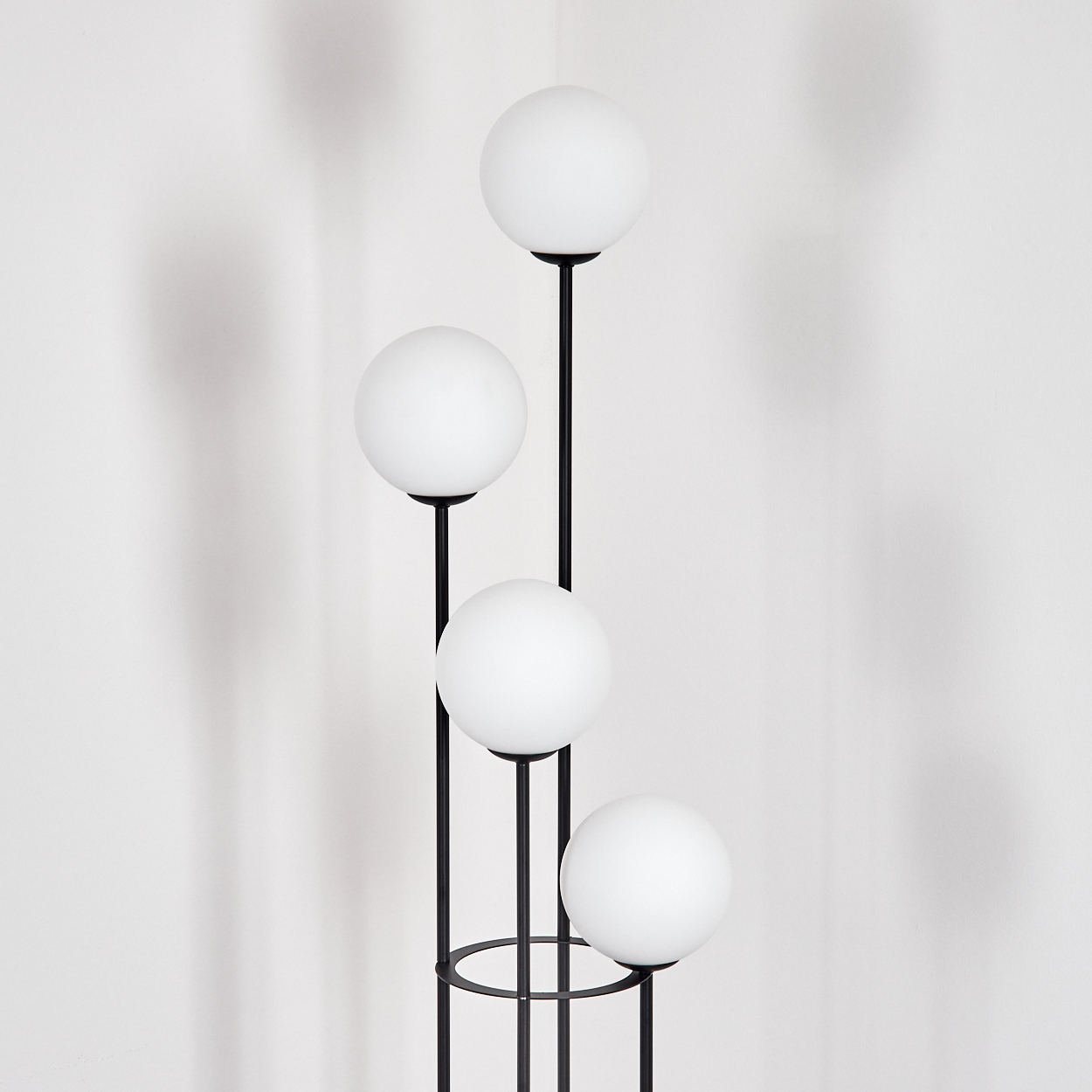 Eine moderne Lampe mit vier weißen Kugelleuchten an schwarzen Metallstielen vor einer weißen Wand. Die Leuchten sind vertikal angeordnet.