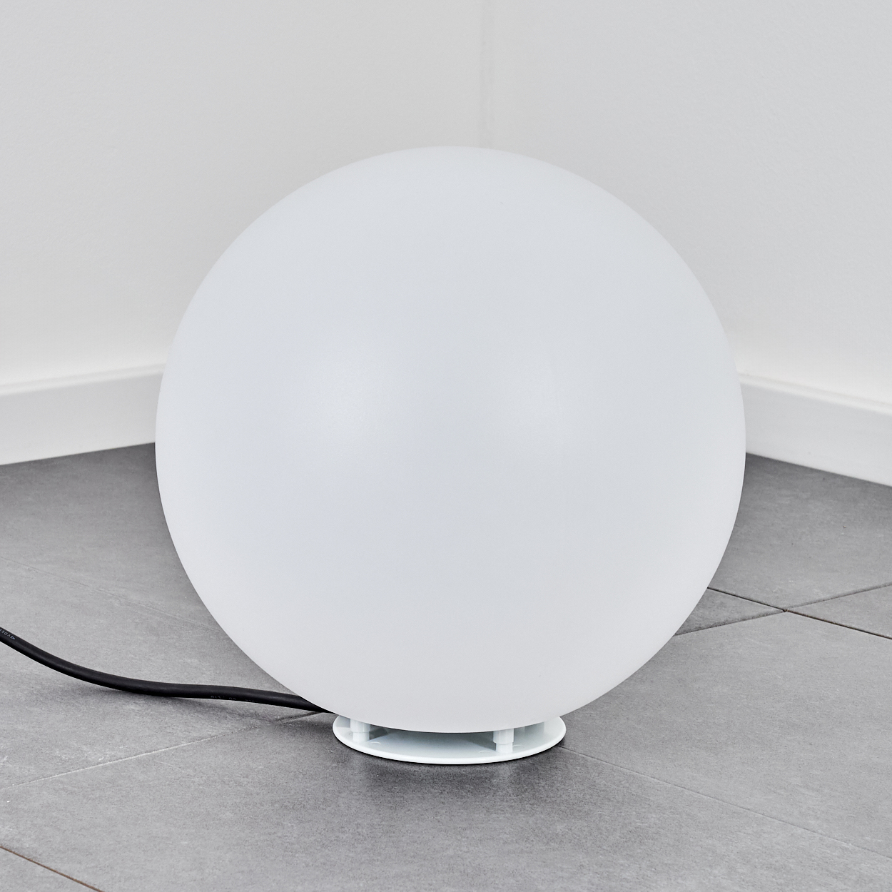 Eine große, weiße kugelförmige Lampe steht auf einem grauen Fliesenboden. Ein schwarzes Netzkabel geht von der Lampe aus.
