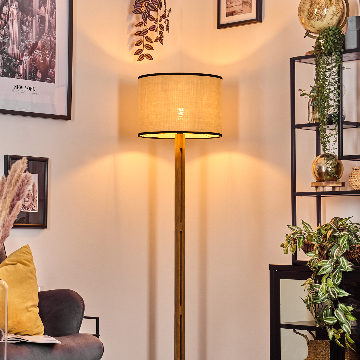 Eine beleuchtete Stehlampe steht in einem Raum mit einem gerahmten New York-Poster, einem Regal mit Pflanzen und einem Sofa mit gelbem Kissen.