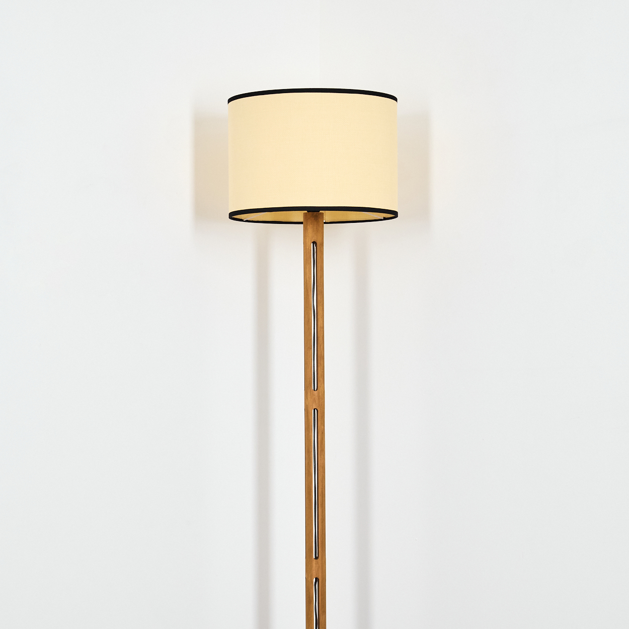 Eine Stehlampe steht vor einer weißen Wand. Sie hat einen beigefarbenen Lampenschirm mit schwarzem Rand und einen Holzpfahl.