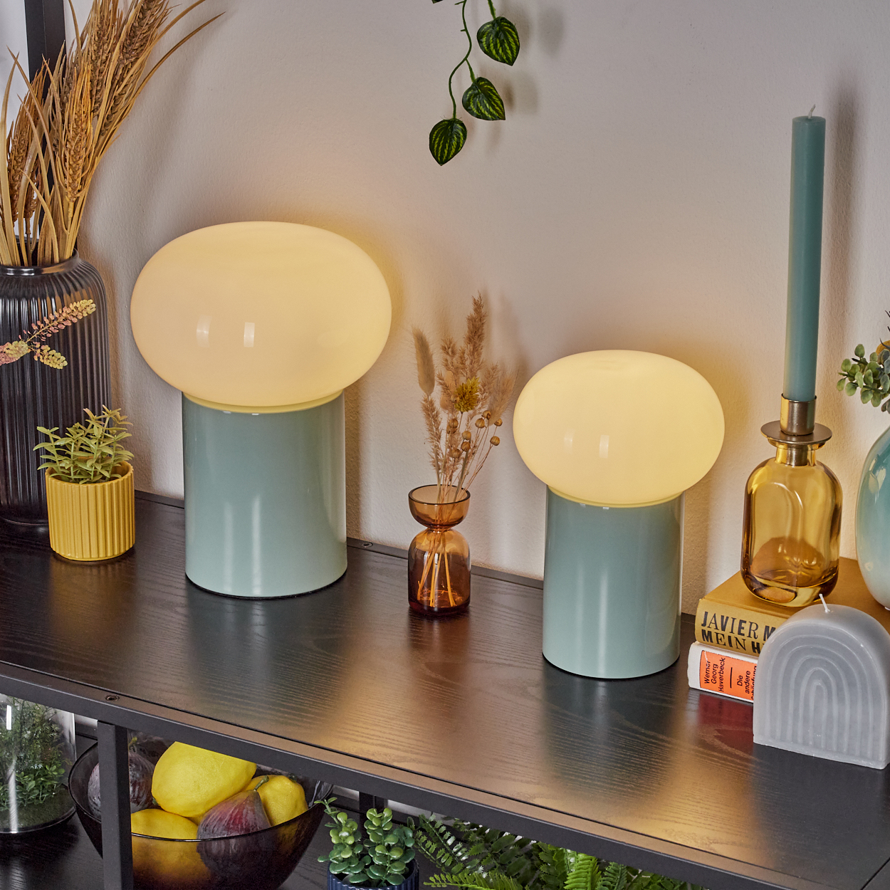 Zwei Pilzlampen auf einem Regal mit Dekoration: Vasen, eine Kerze und Pflanzen. Früchte in einer Schüssel darunter. Ruhige, moderne Innenszene.