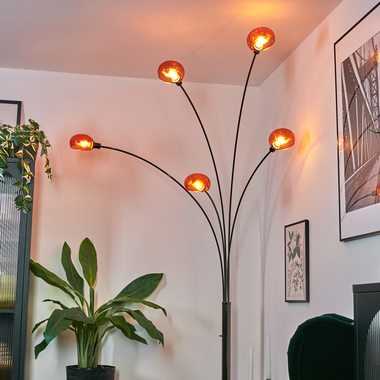 Eine moderne Stehlampe mit fünf orangefarbenen Glaskugeln, schwarzem Rahmen und einer großen Topfpflanze. Der Hintergrund umfasst Wandkunst.