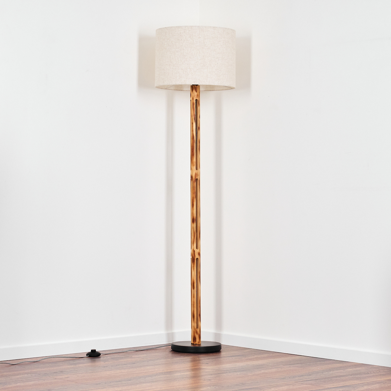 Eine hohe Stehlampe steht in einer Ecke. Sie hat einen beigen Lampenschirm, einen bambusartigen Holzpfahl und einen schwarzen Sockel.