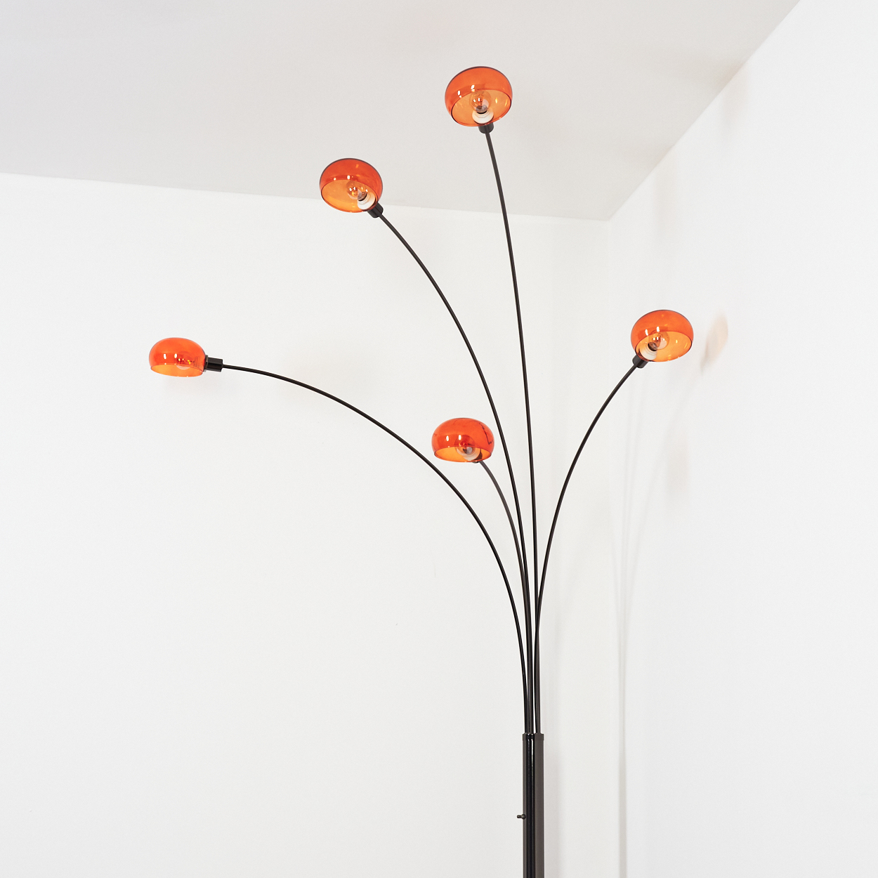 Eine moderne Stehlampe mit fünf orangefarbenen Glasschirmen. Der schwarze Metallrahmen ist gebogen. Der Hintergrund ist eine weiße Wand.
