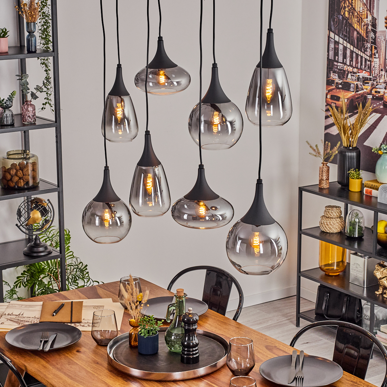 Esszimmer mit Holztisch, schwarzen Stühlen und Hängelampen. Tisch gedeckt mit Tellern und Gläsern. Regale mit Dekoration.
