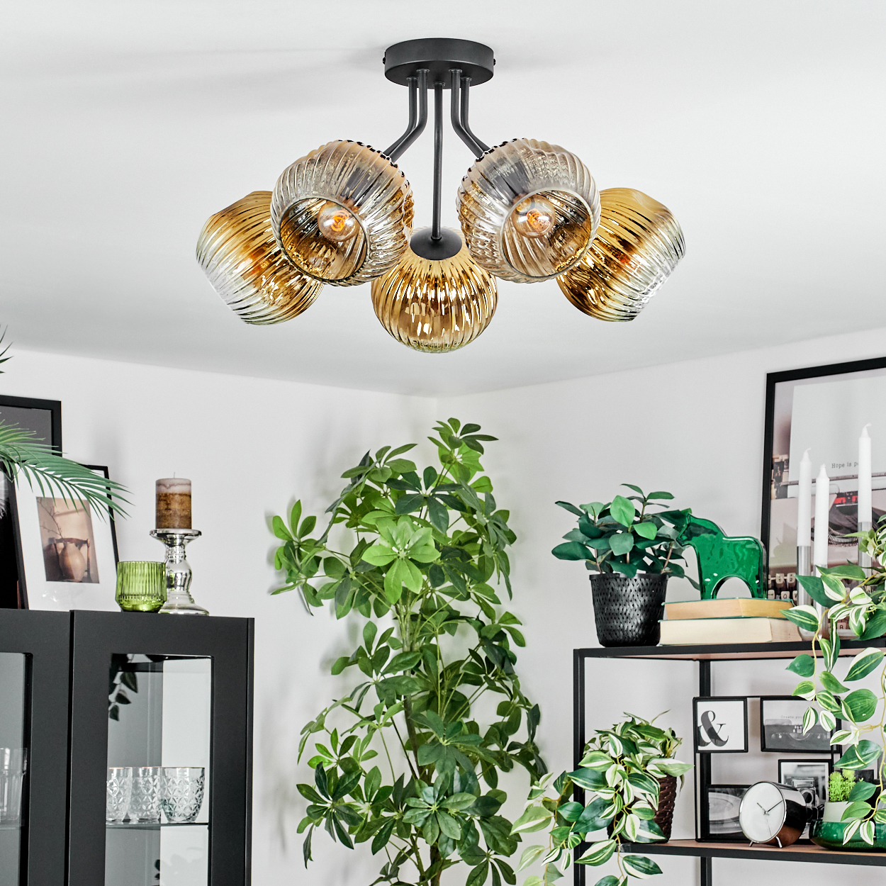 Deckenlampe mit Glasschirmen, Raum mit Pflanzen, Kerzen und gerahmten Bildern. Schwarzes Regal mit Deko und einem grünen Pferd.
